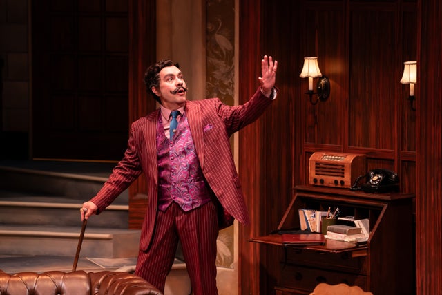 An actor in a red suit with a distinctive mustache poses onstage