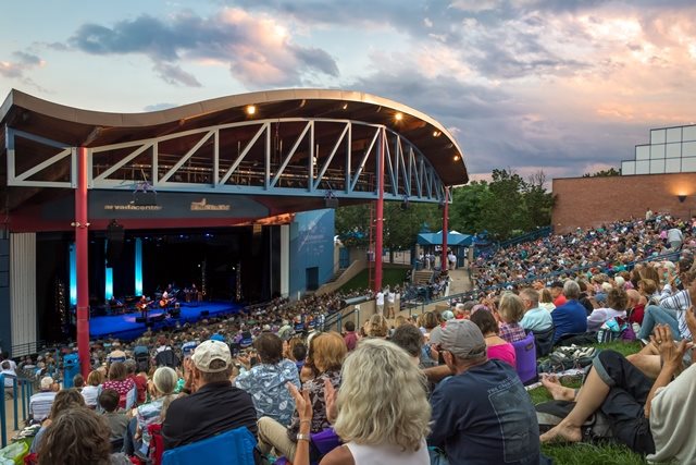 A crowd of concert attendees look at the Arvada Center's outdoor stage, with the sun setting in the background.