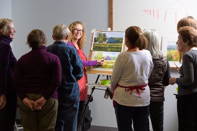 A teachers shows a watercolor painting to a group of adult students, who look on with interest.